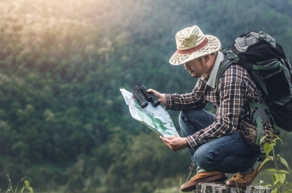 Man with hat and backpack squatting down in the mountains looking at a map.